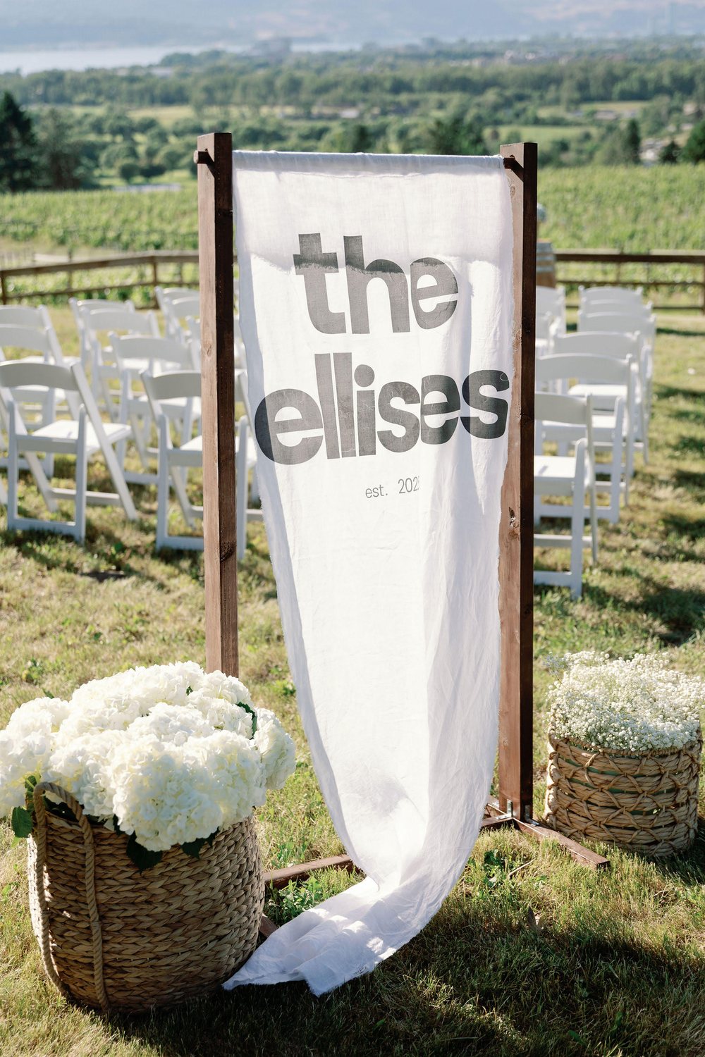 Linen ceremony banner on a wooden frame flanked by woven baskets of white hydrangeas and baby's breath, with white chairs and vineyard hills behind