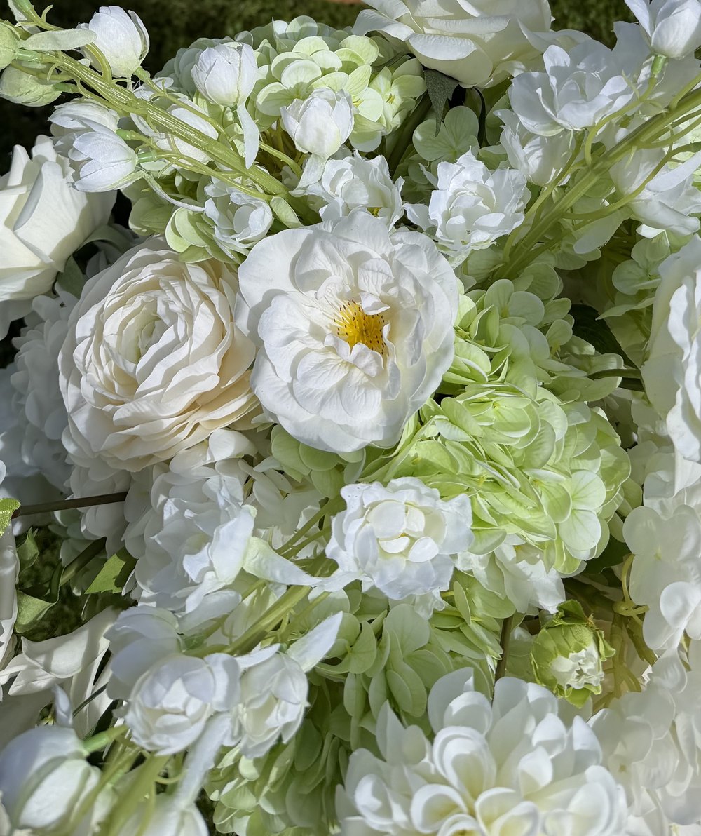 Close up of a real-touch arrangement: ivory garden roses, an open white rose with a golden centre, cream ranunculus, pale green hydrangea and white delphinium sprays in bright sun