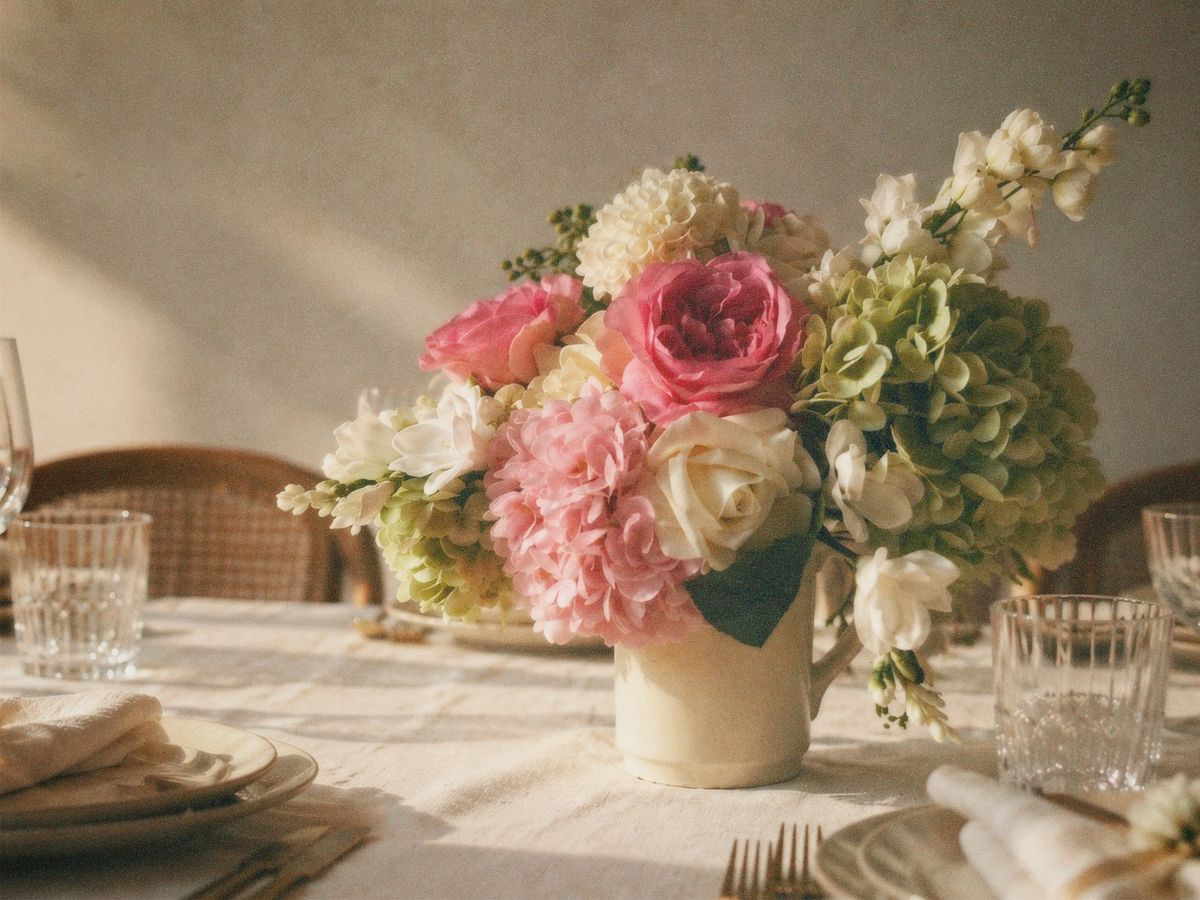 Centrepiece of pink garden roses, blush dahlias, cream roses, white stock and green hydrangea in a cream pitcher on a linen-set table in warm light